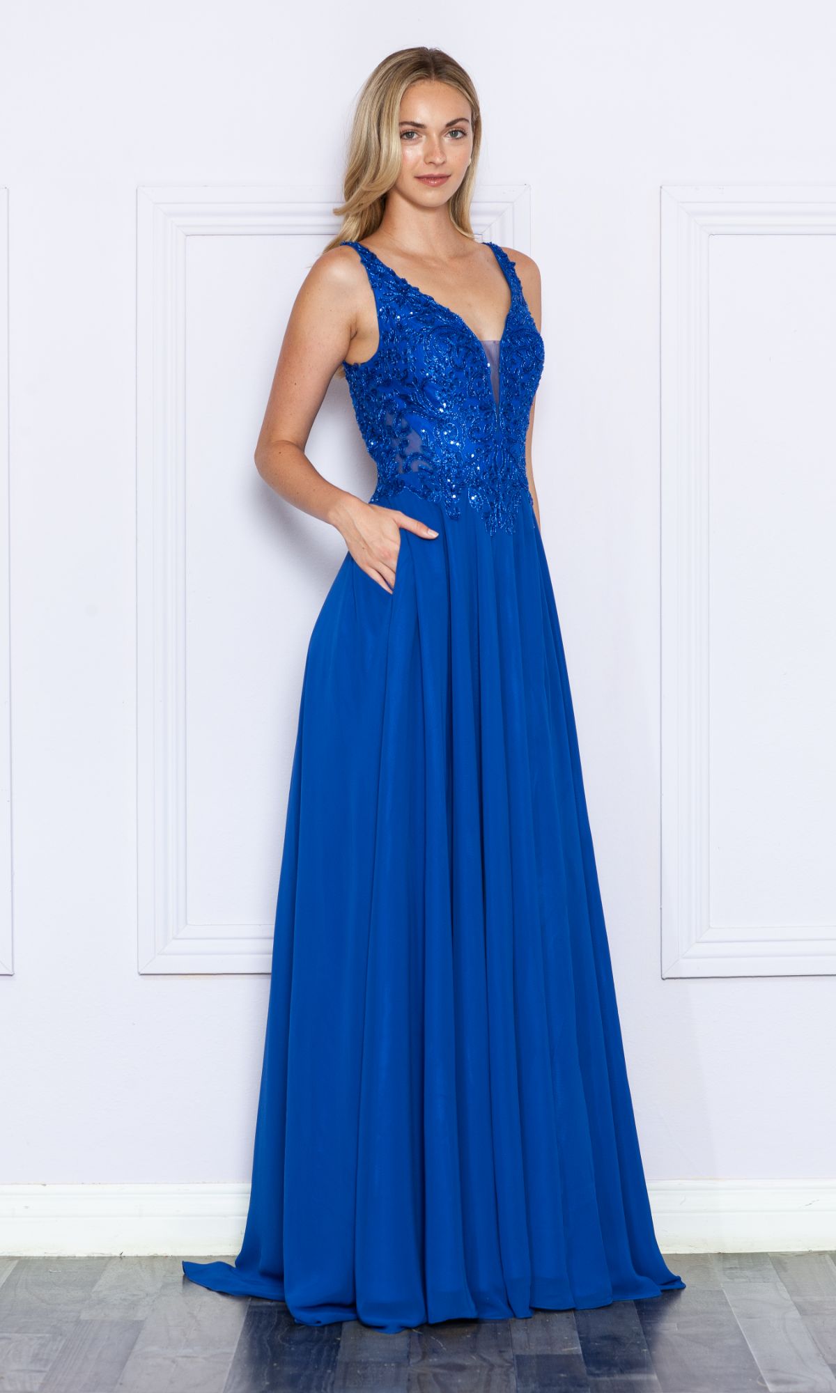 A woman stands against a white wall wearing the Beaded-Bodice Long A-Line Prom Dress 9366, featuring a sparkly beaded bodice and a flowing blue skirt.